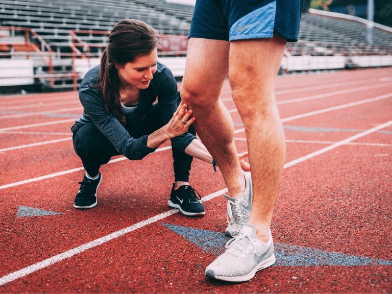 female running coach is kneeling on a track while physically moving a runner's leg from the knee and to the ankle of the runner's right leg with both of her hands