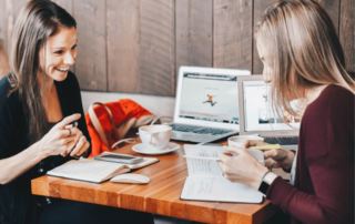 two women sitting at a cafe table comparing notes and laughing