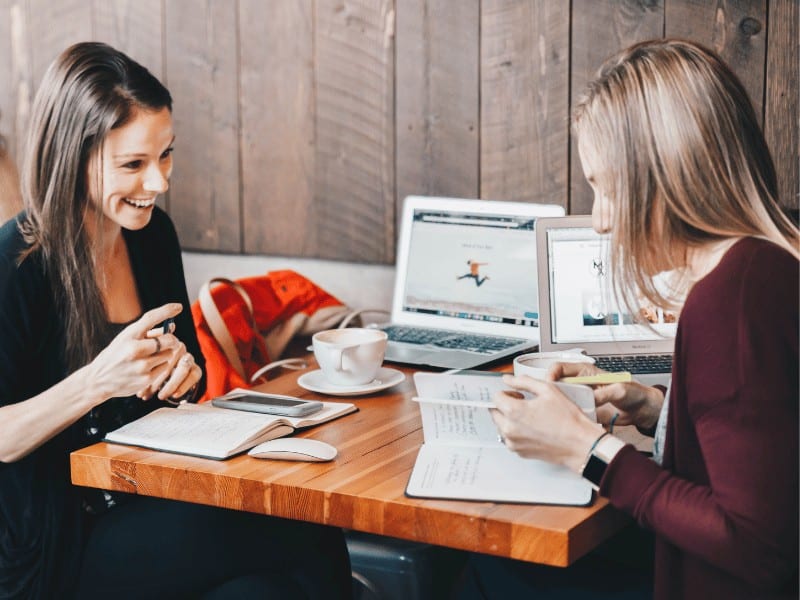 two women sitting at a cafe table comparing notes and laughing