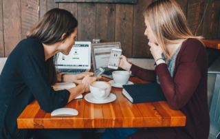 two women sitting at a cafe table are comparing how an image looks on a mobile device versus a laptop screen