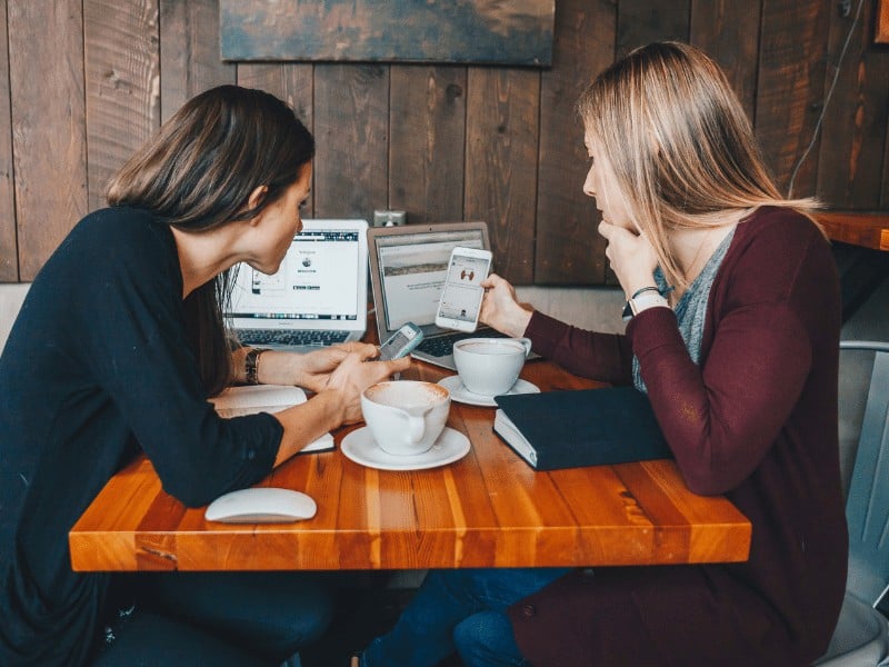 two women sitting at a cafe table are comparing how an image looks on a mobile device versus a laptop screen