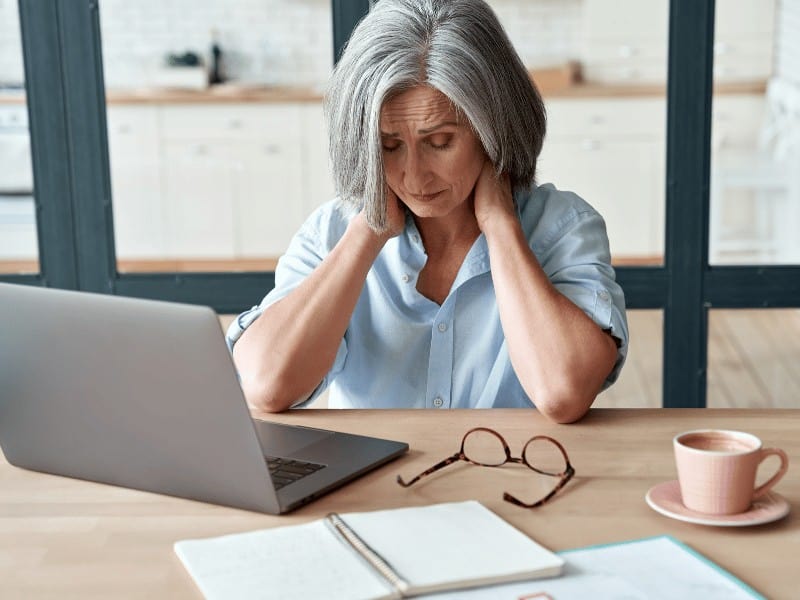 Senior woman experiencing chronic pain sitting at her desk grabbing her neck in discomfort
