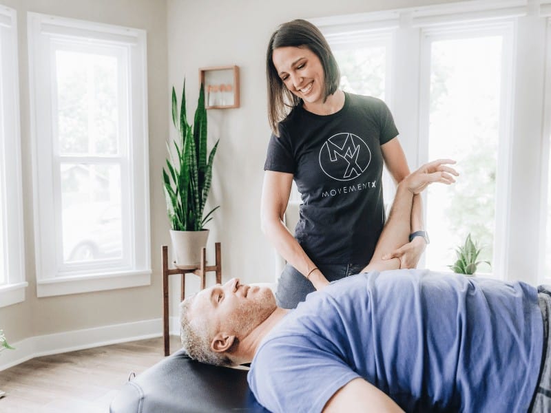 physical therapist with movementx performing a hands-on manual therapy technique on a male patient with shoulder pain on a portable treatment table in his bright sunny living room
