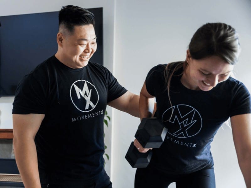 MovementX physical therapy in portland provider Dr. Steven Park wearing a black movementx physical therapist shirt smiling while helping a female patient get stronger during a physical therapy appointment at home by lifting weights with her shoulder via a bent over row exercise