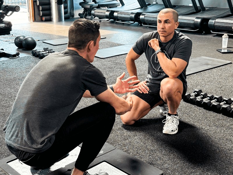 Derek Garza and a patient in a squat position having a discussion in a CrossFit gym workout area.