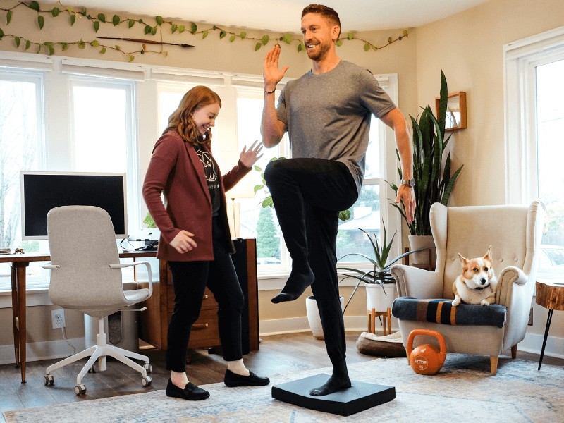 A MovementX provider watching a patient perform standing one leg balance exercises on a foam pad in their living room.