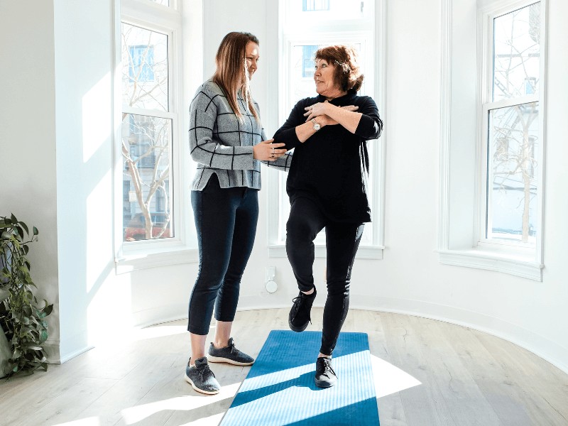 A MovementX provider helping a patient perform standing balance drills on one leg in an open front room at their house.