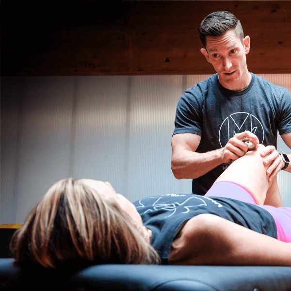 A male clinician providing physical therapy for athletes via myofascial release to a female patient's knee in a clinic setting.