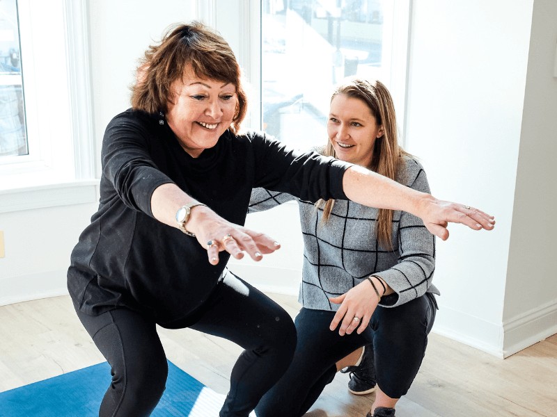 A movementx physical therapy provider instructing an older woman to squat with proper mechanics to avoid arthritis pain in a bright home setting.