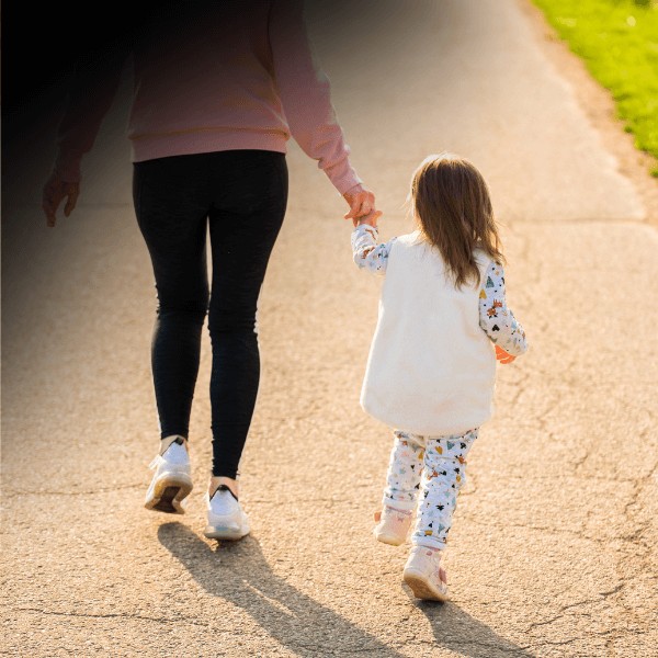 A mother holding her daughter by the hand and walking away down a bright sidewalk.
