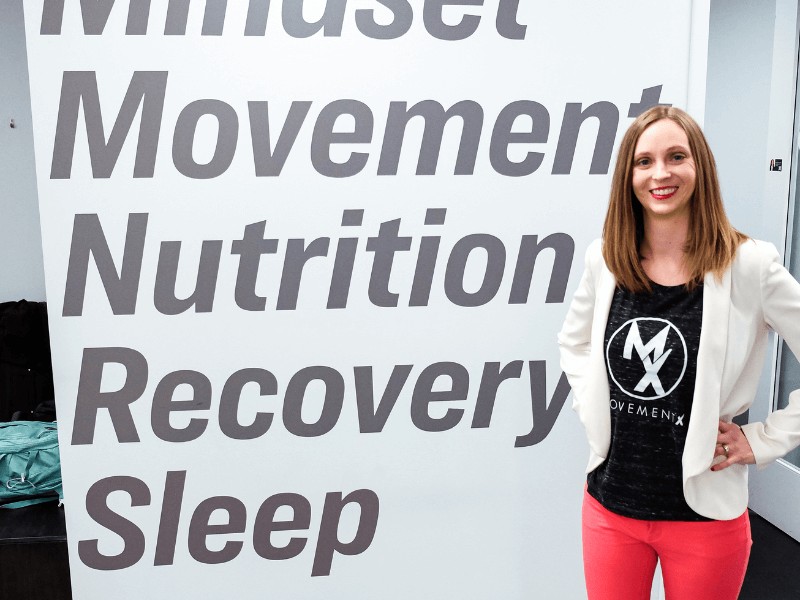 A movementx pelvic health physical therapist standing next to a clinic wall with the words Movement, Nutrition, Recovery, and Sleep written on it.