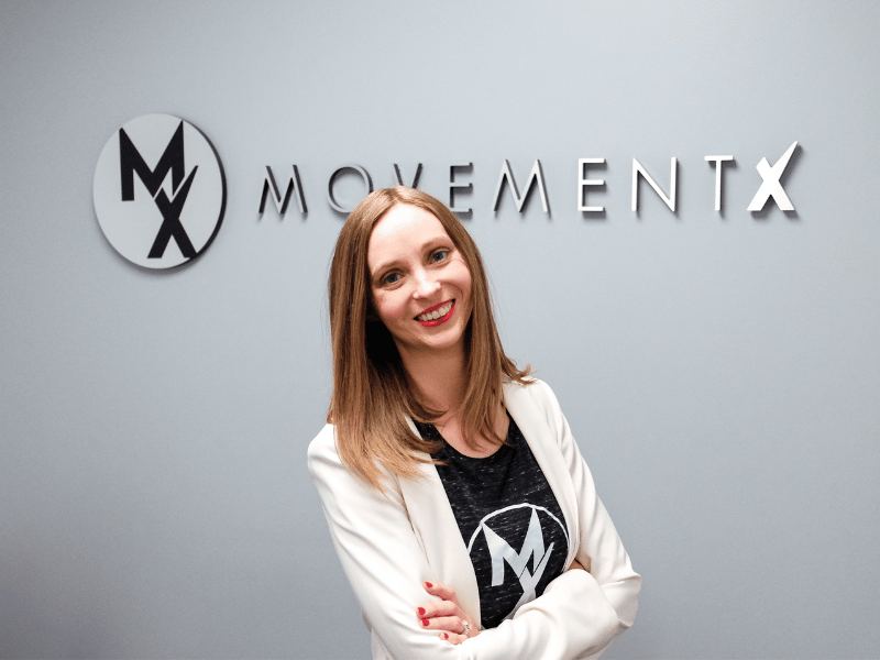 Dr. Stephanie Weyrauch, PT, DPT standing in front of a MovementX logo on her treatment area's wall.