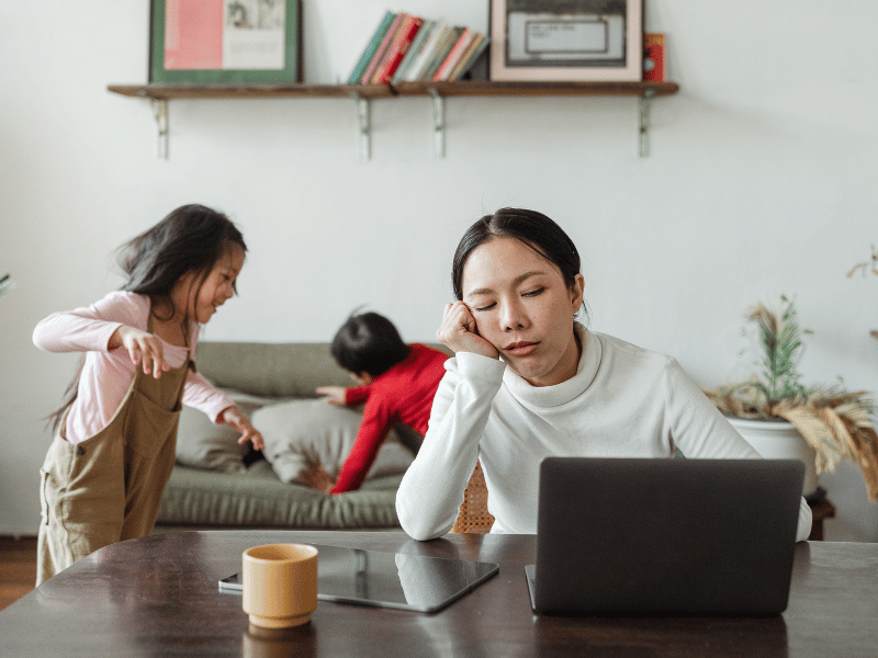 A tired woman sitting in front of her laptop during the day with her children playing behind her.