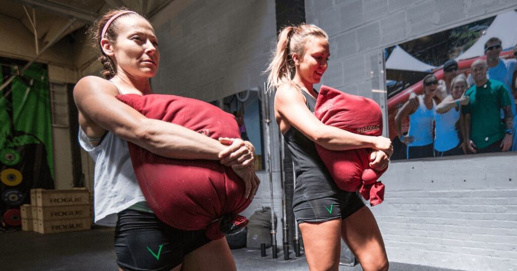 Two women in a gym front carrying a large weighted red sandbag