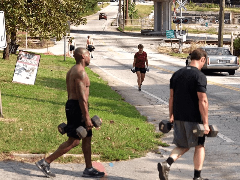 A workout class doing farmer's walks and carries down a street.