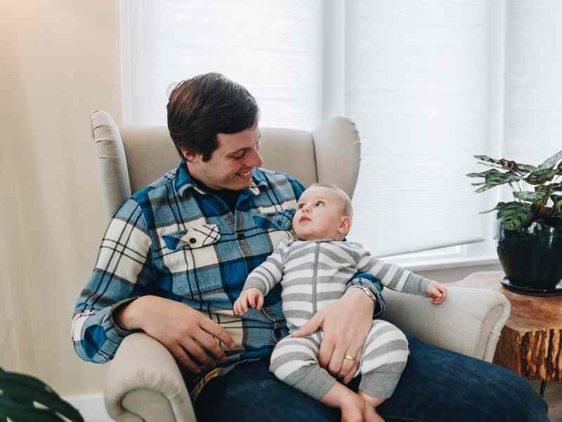 A new parent sitting on an armchair, holding his baby while looking down at him.