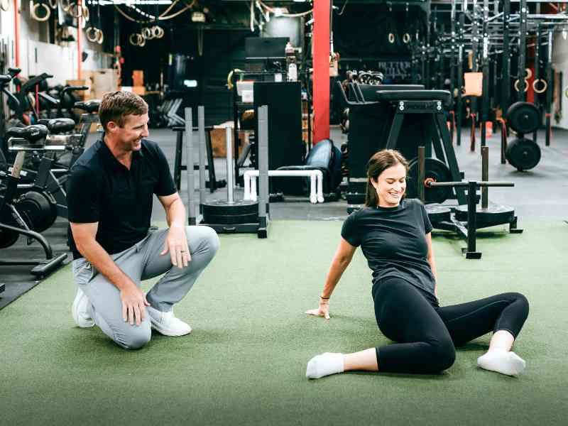 MovementX provider Tyler Eubanks, PT, DPT helping a patient perform 90-90 hip opener stretches on turf in a gym.