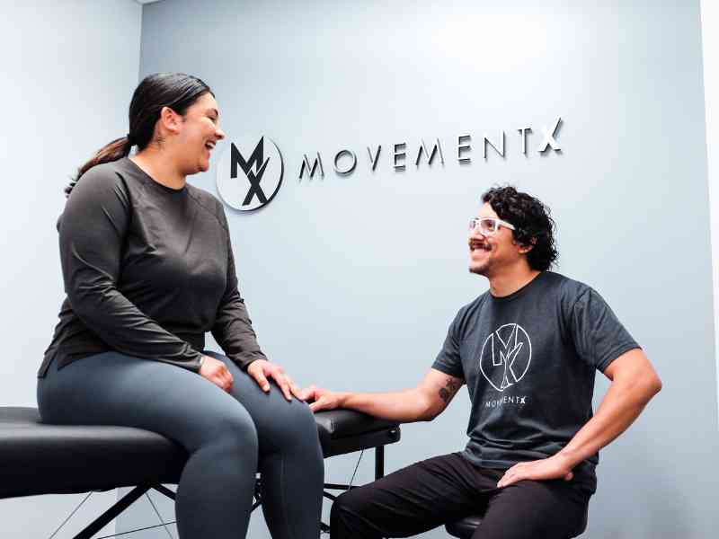 Rich Ortiz, PT, DPT sitting on a stool in a private treatment room smiling and talking with a patient who is sitting on a treatment table during an initial consultation in Washington, D.C.