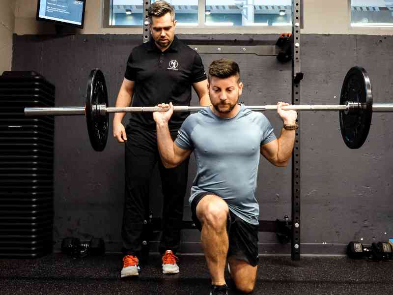James Cousler, PT, DPT helping a patient do a split squat with a barbell during a session in a gym.