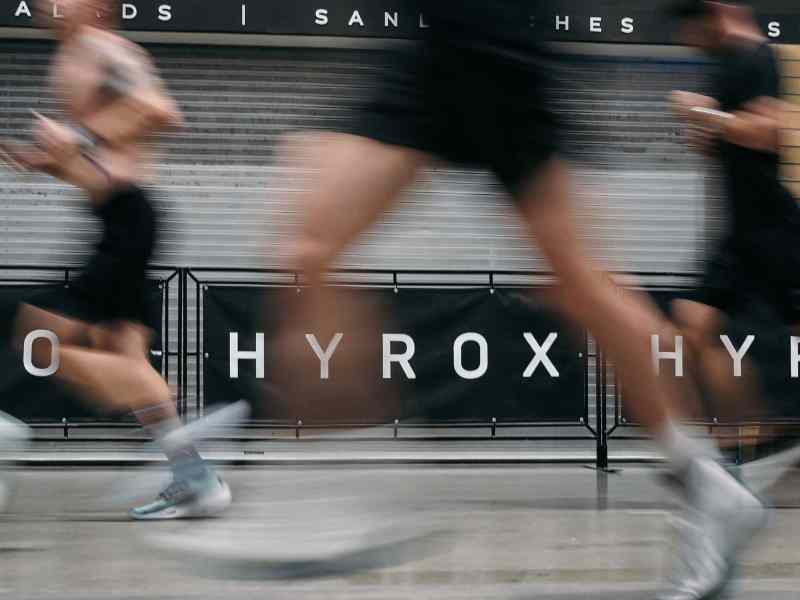 A group of athletes running past a Hyrox sign indoors.