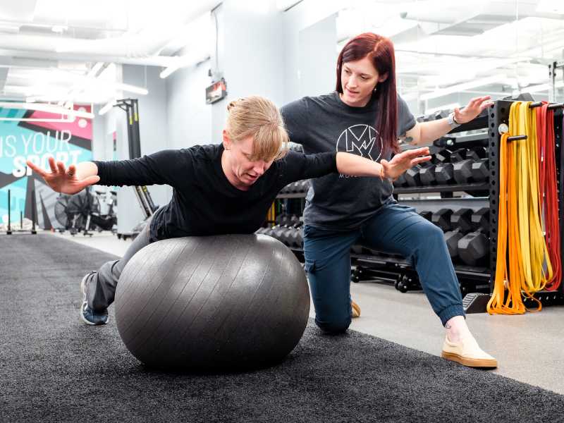 MovementX provider Colleen Gallagher, PT, DPT helping an older patient do a balance exercise on a bosu ball in a gym treatment session