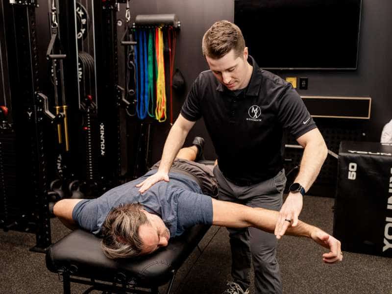 MovementX Provider Adam Kays helping a patient assess shoulder mobility on a treatment table in a gym.