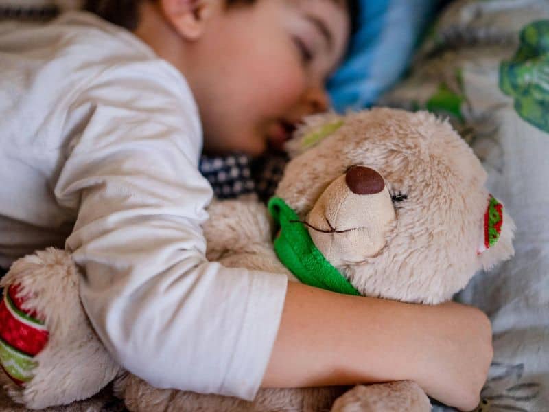 A child sleeping with a teddy bear in bed.